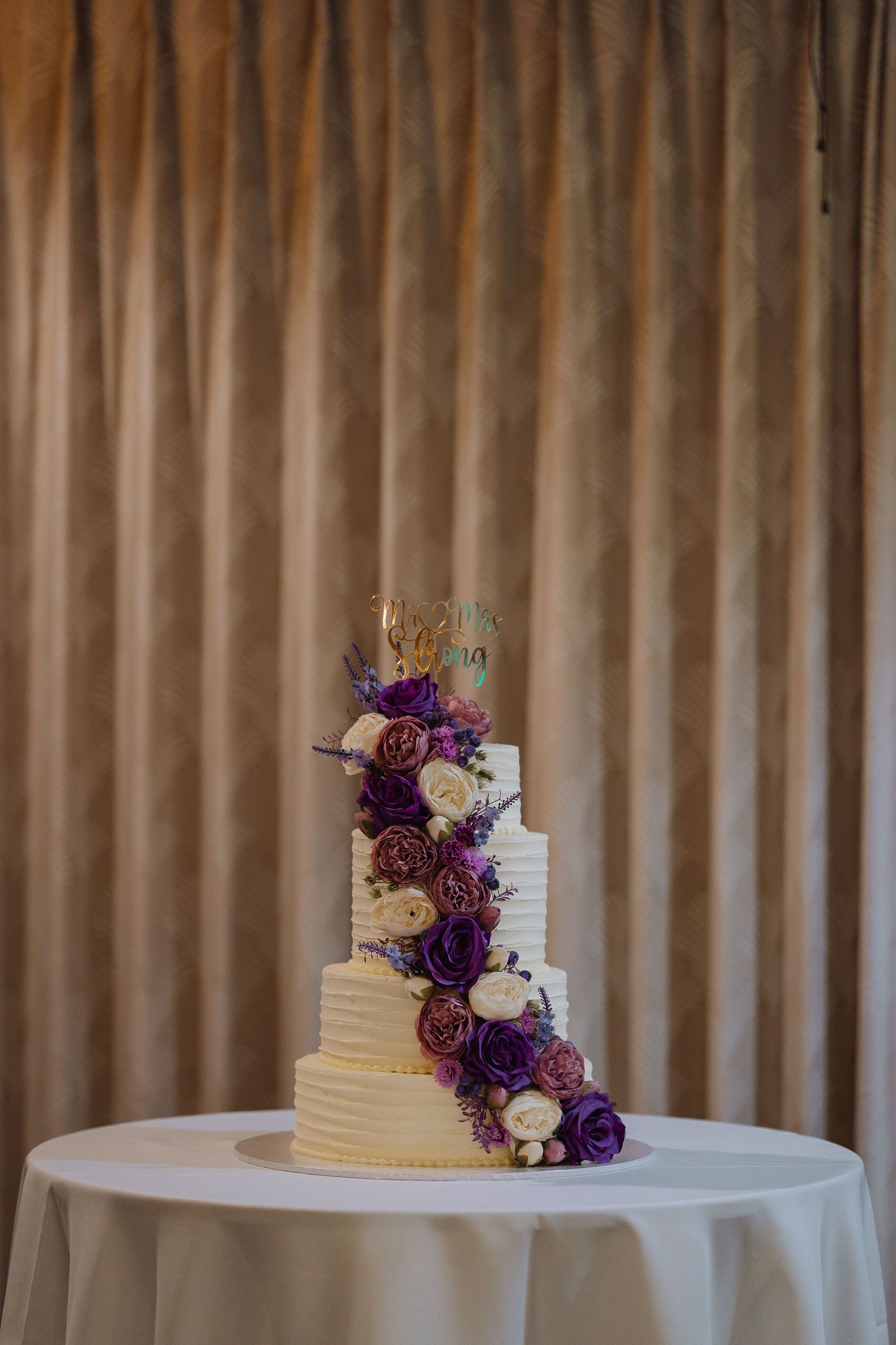 Elegant white wedding cake decorated with cascading purple and burgundy flowers on a round table.