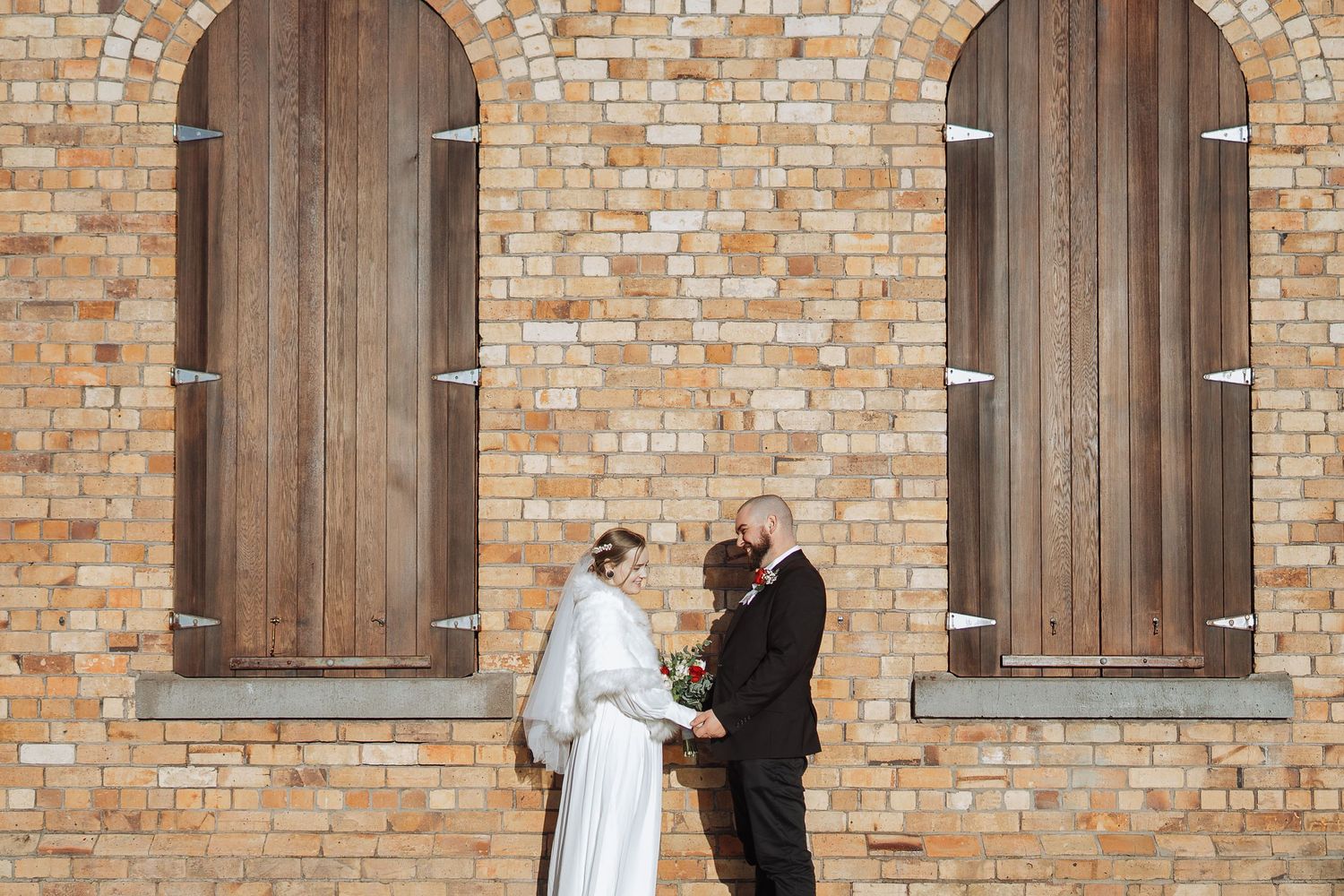 Wedding couple poses against rustic brick wall with wooden doors.