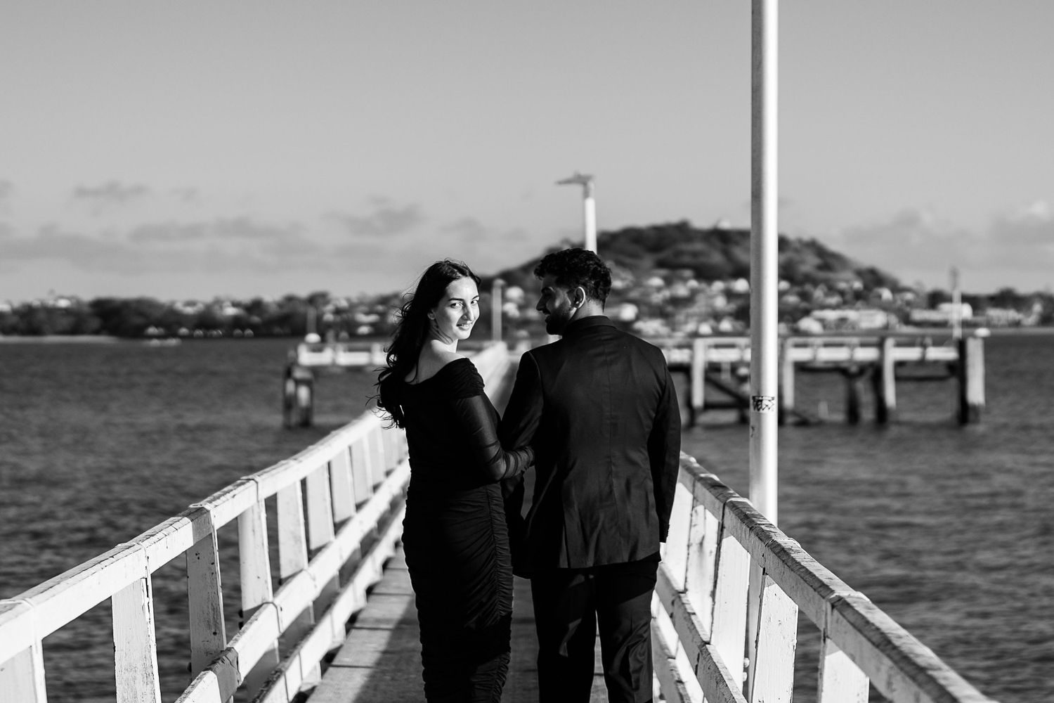A couple stands together on a white pier overlooking the bay.