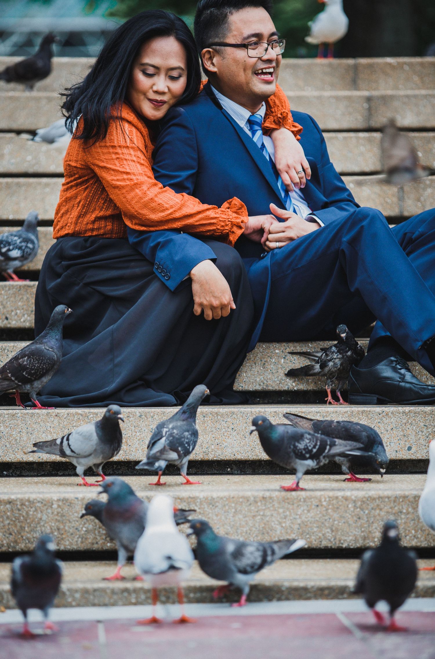 Couple sitting on steps surrounded by pigeons, woman in orange top and man in blue suit smiling together outdoors.