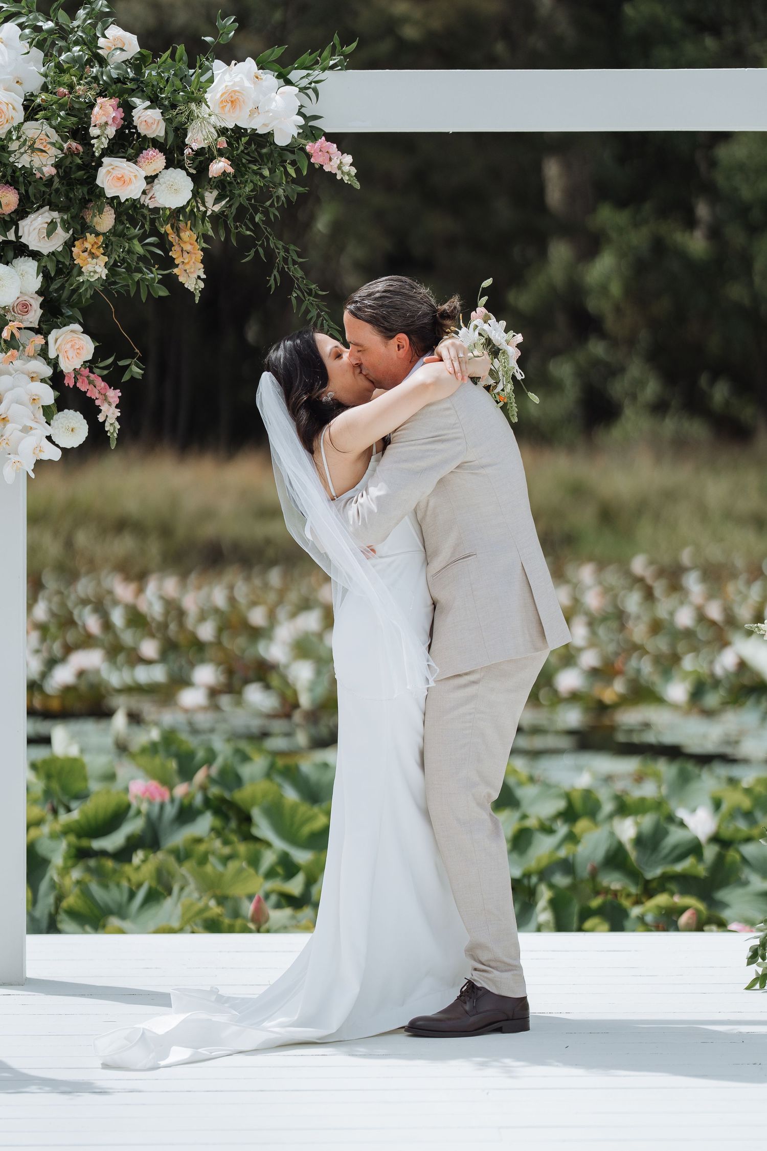 Newlyweds share their first kiss under a white arch decorated with peach and white flowers.