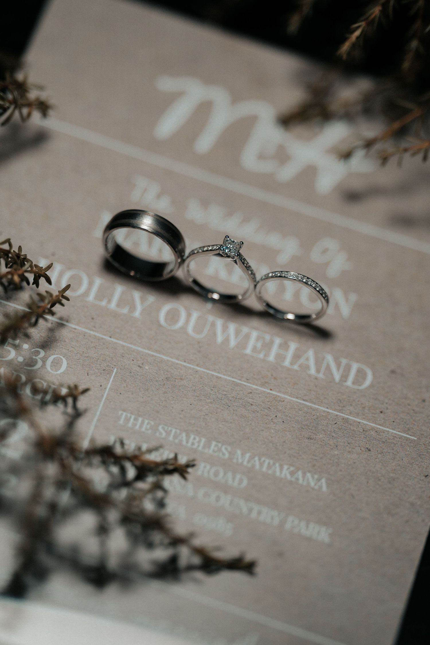 Wedding rings and invitation details photographed on a beige textured surface with delicate floral elements.