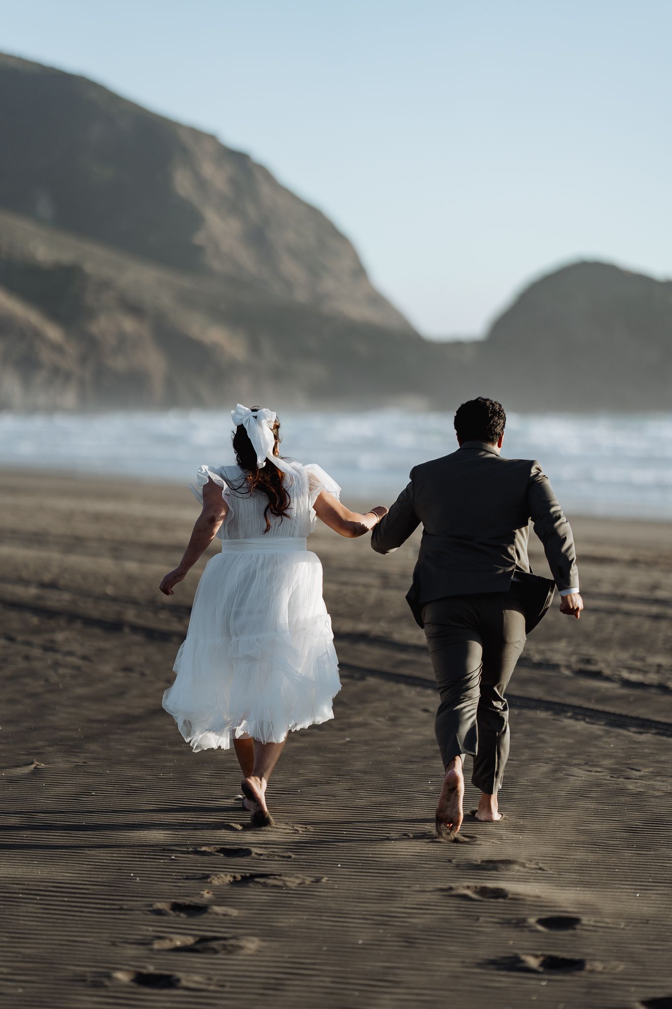 Couple runs joyfully across dark sandy beach with coastal cliffs in distance.