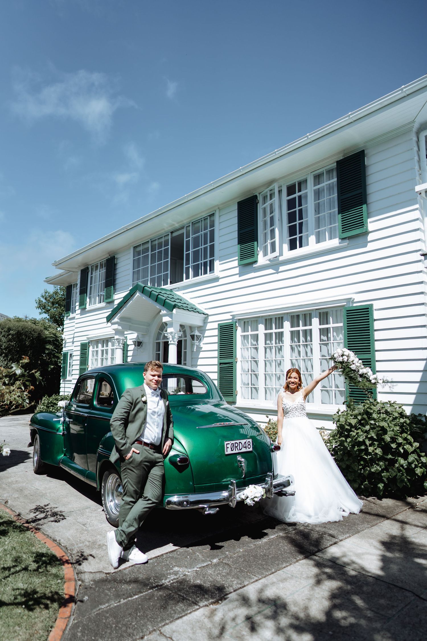 Wedding couple poses with vintage green car outside white colonial-style house.