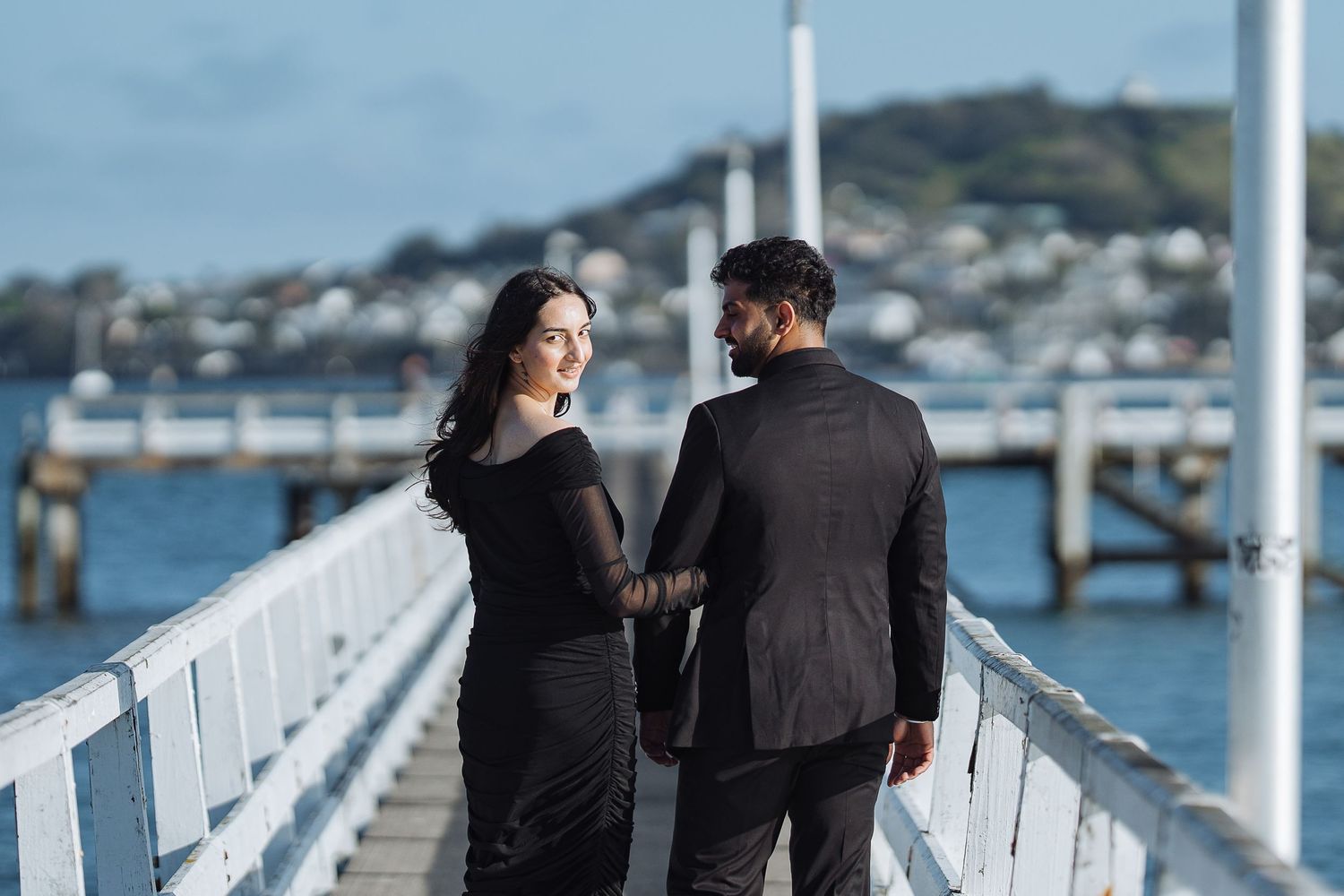 A couple walks together on a white pier overlooking a waterfront on a sunny day.