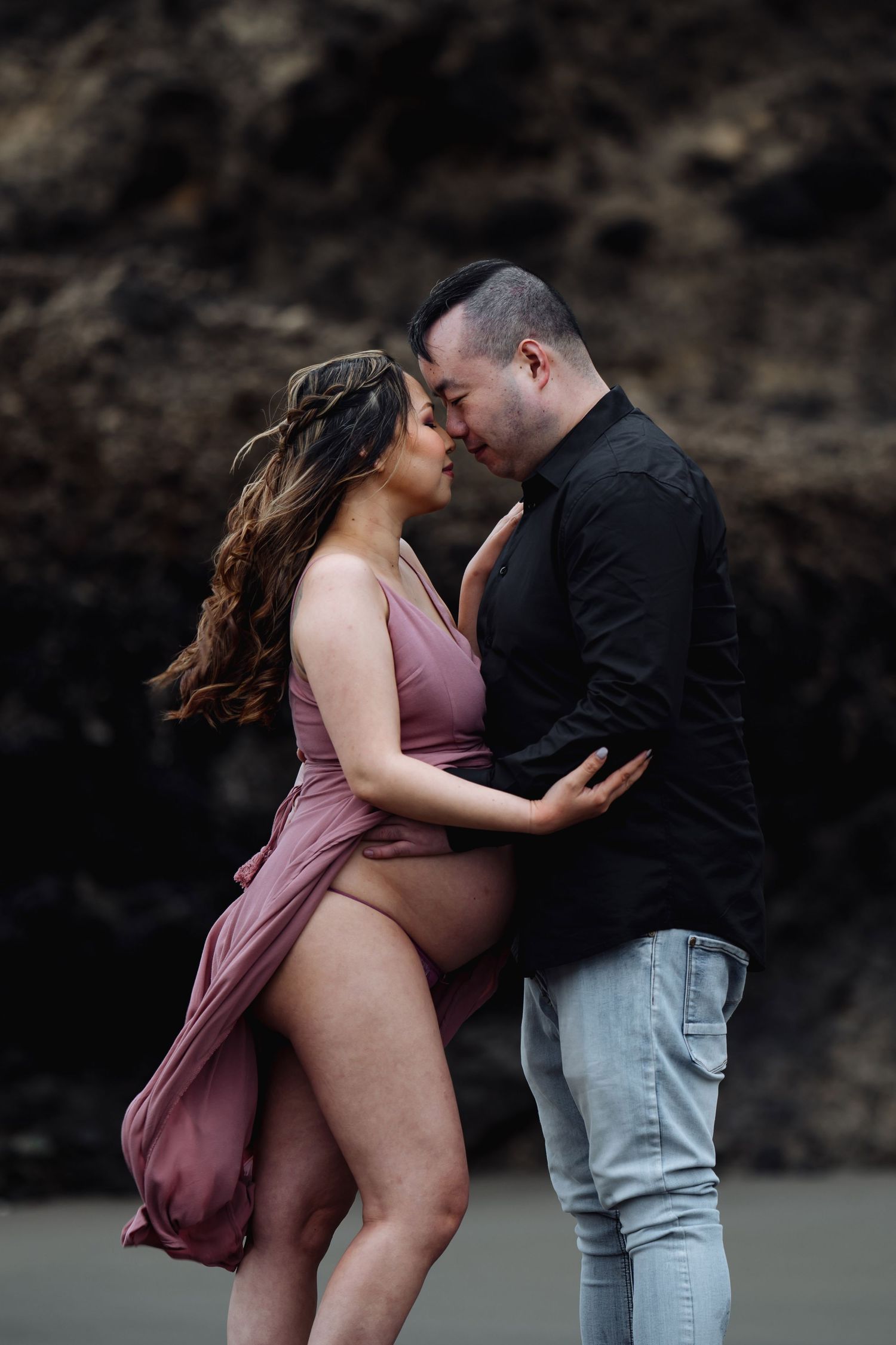 A romantic couple embraces on a beach with dramatic rocky cliffs in the background.