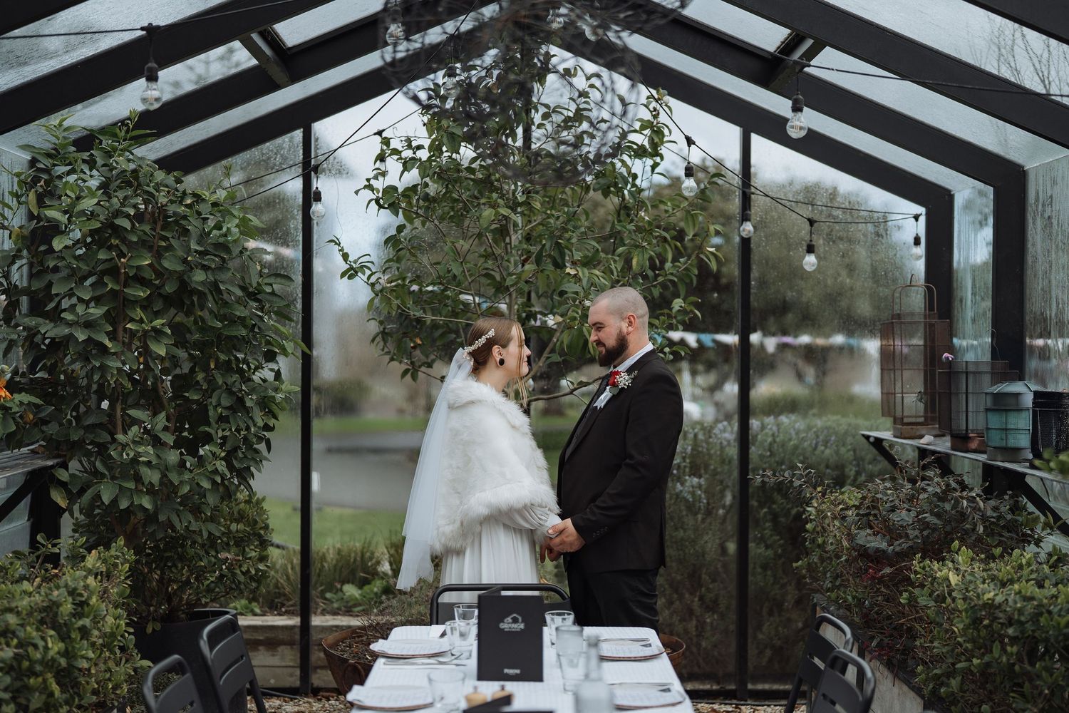 Wedding reception setup in modern glass conservatory with hanging lights.