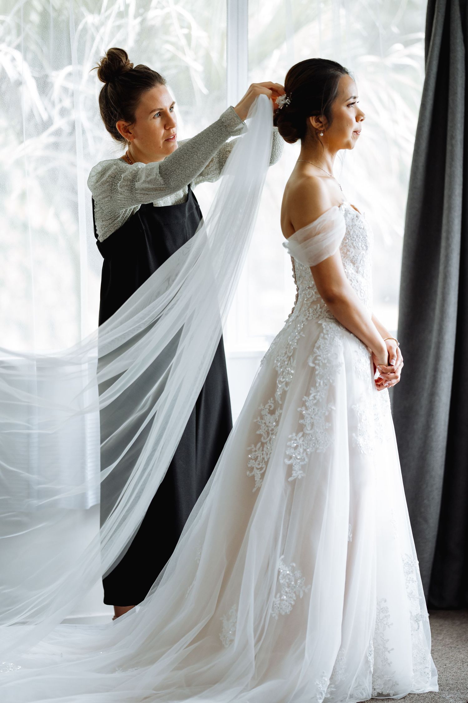 A wedding dress with lace details and flowing veil being adjusted in front of bright windows.