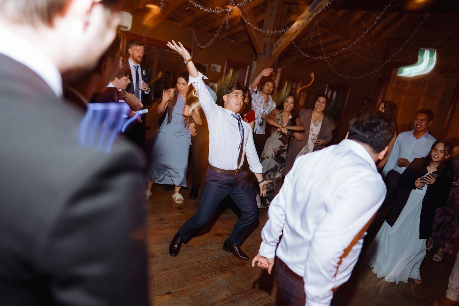 Wedding guests dance and celebrate in formal attire under atmospheric lighting at an indoor venue.