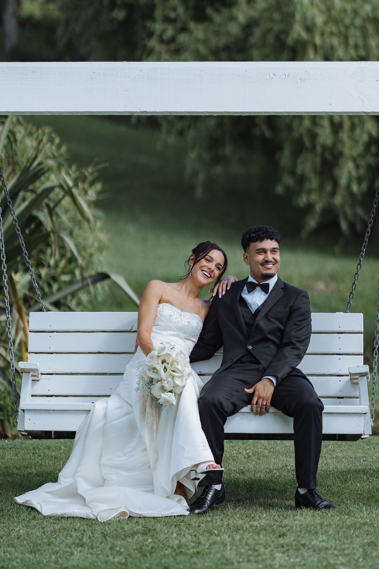 Couple sitting together on white bench in garden setting wearing formal wedding attire.