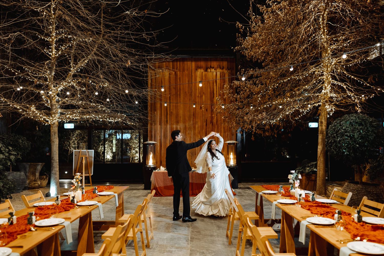 Long wooden tables decorated with orange accents under illuminated bare trees at a night wedding reception.