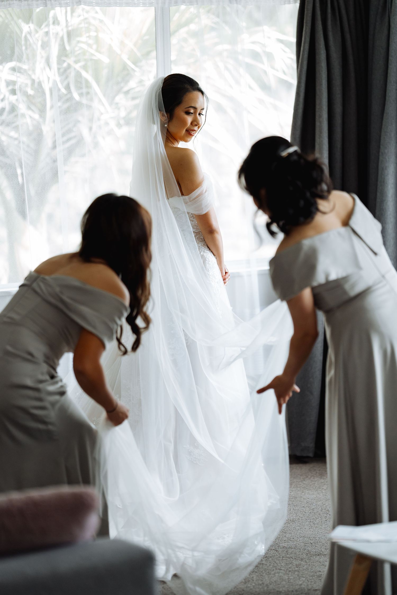 Bridesmaids help adjust a wedding dress and veil in a bright room with white curtains.
