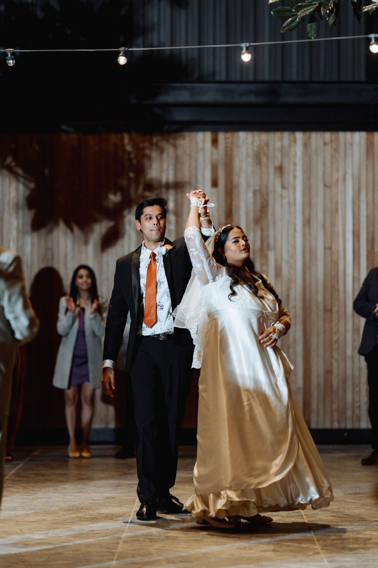 Couple dancing at wedding reception with string lights and wooden backdrop.