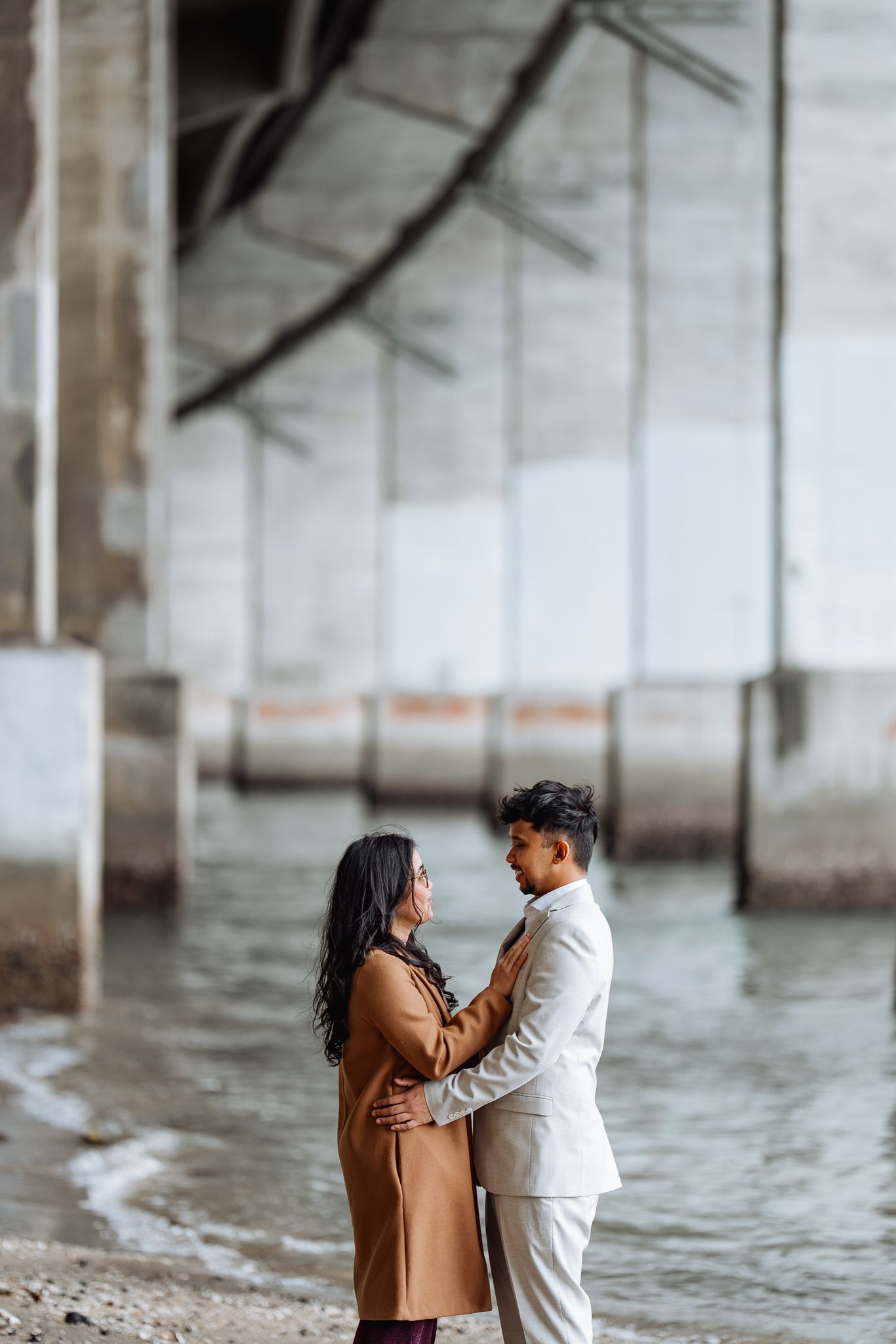 Couple embracing under an industrial structure near water during an overcast day.