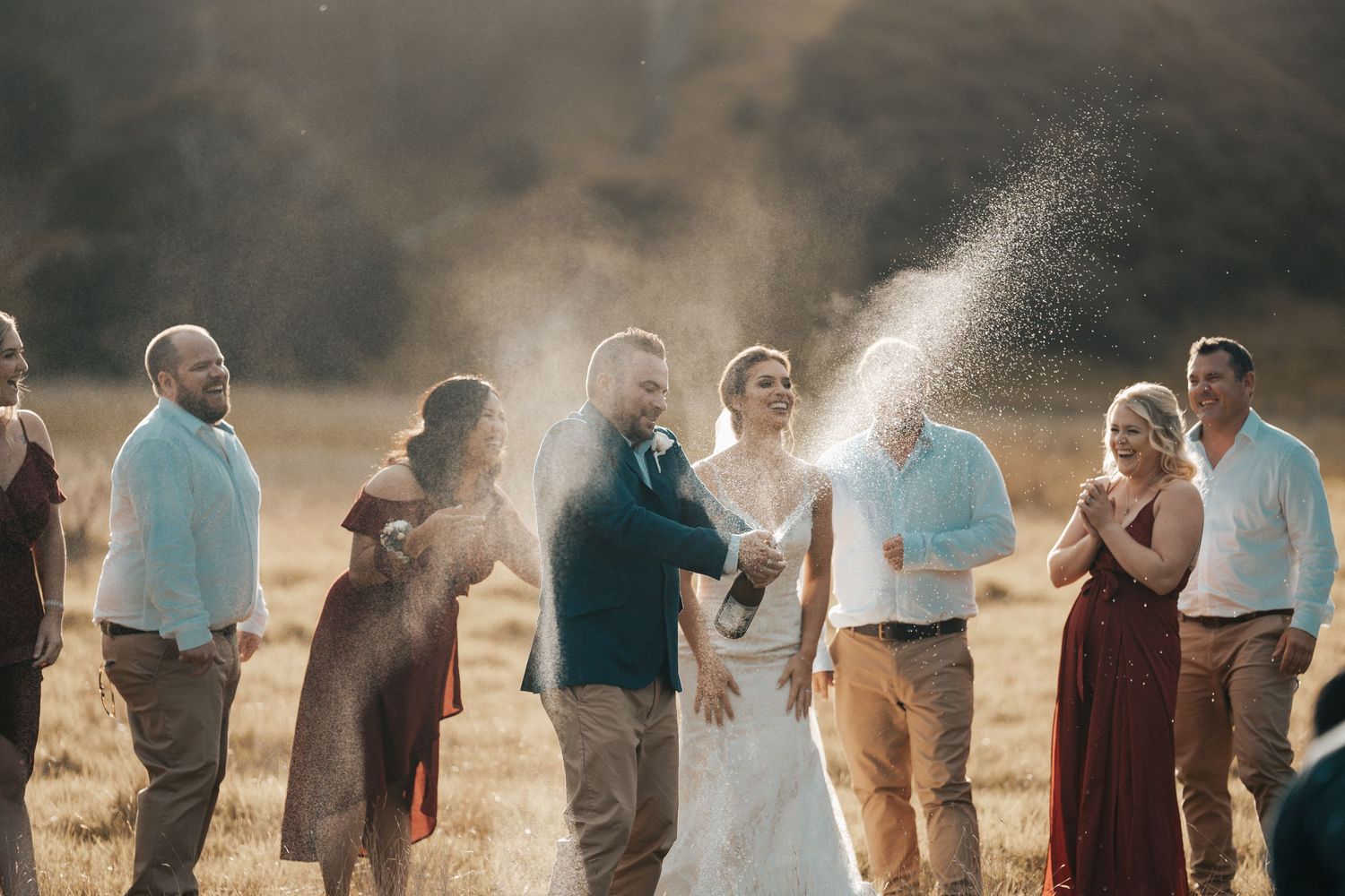 Wedding celebration in golden field with champagne spray and joyful guests.