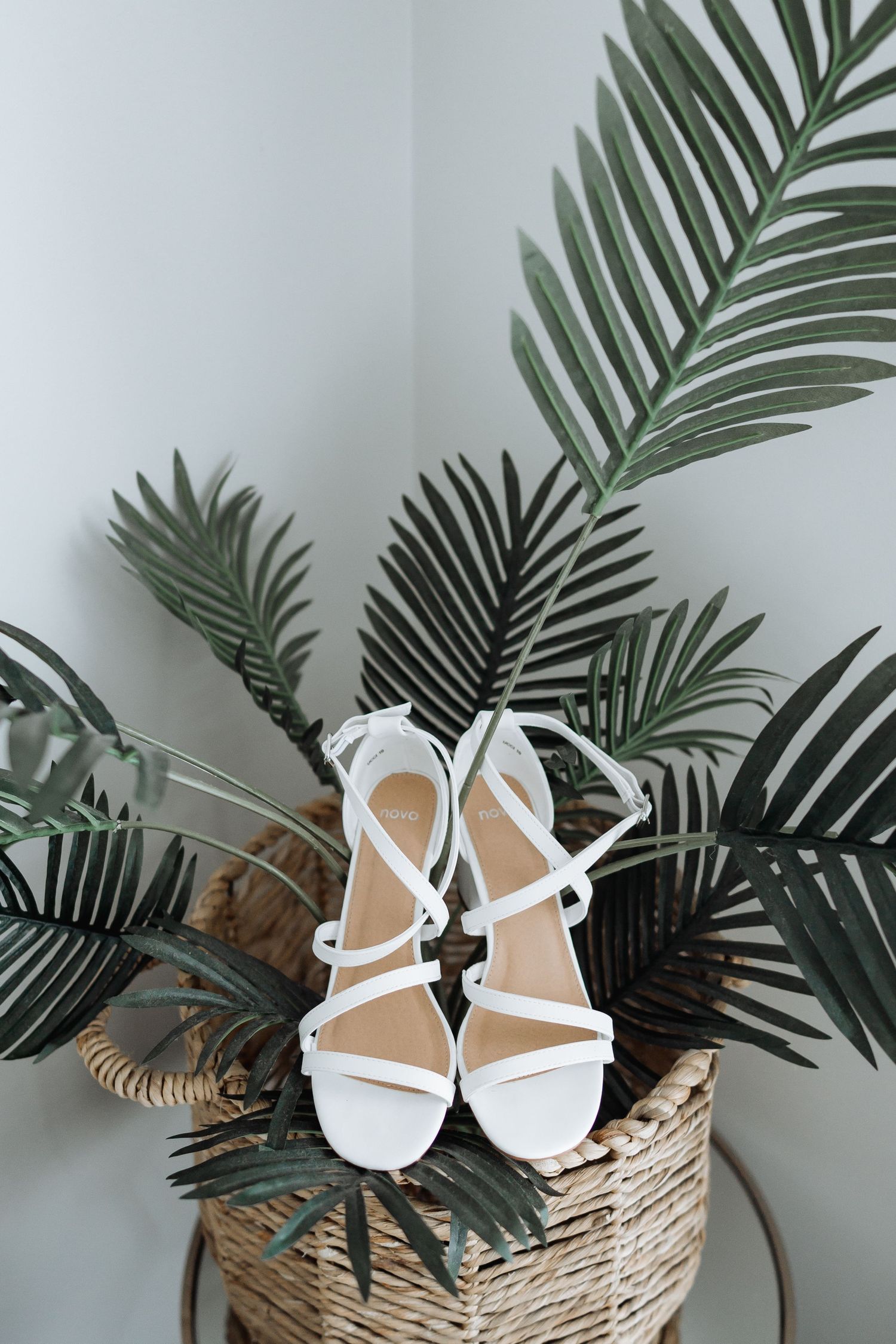 White bridal sandals displayed with tropical palm leaves on a woven basket.