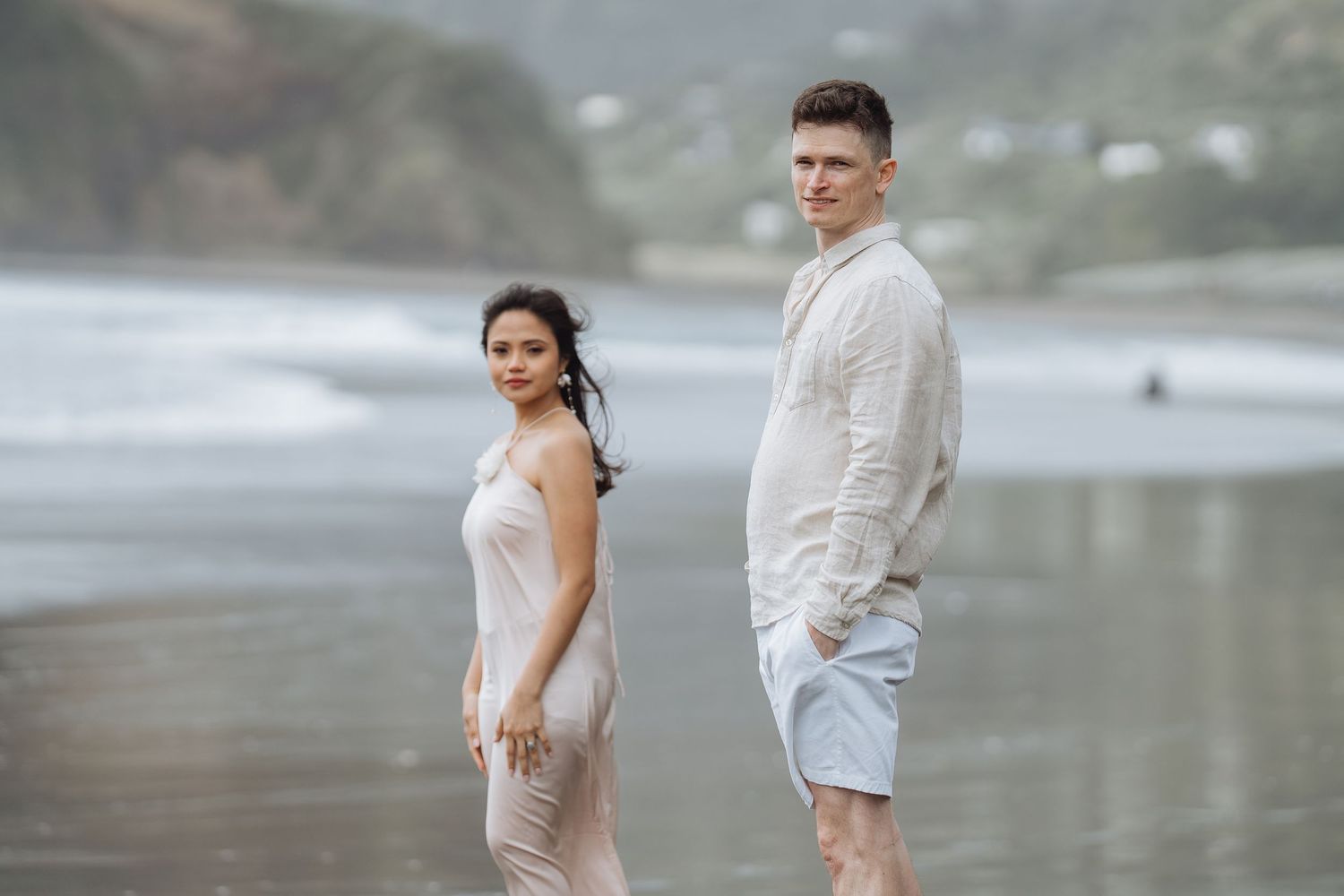 Couple in white clothing walking along misty beach with mountains in background.