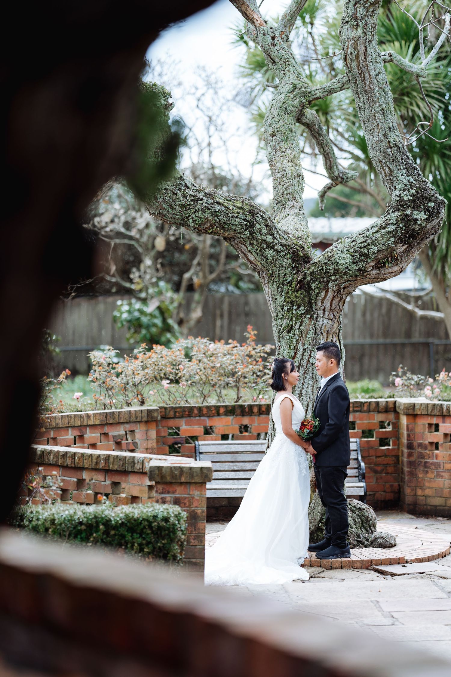 Wedding couple poses on rustic brick pathway surrounded by overhanging trees and garden scenery.