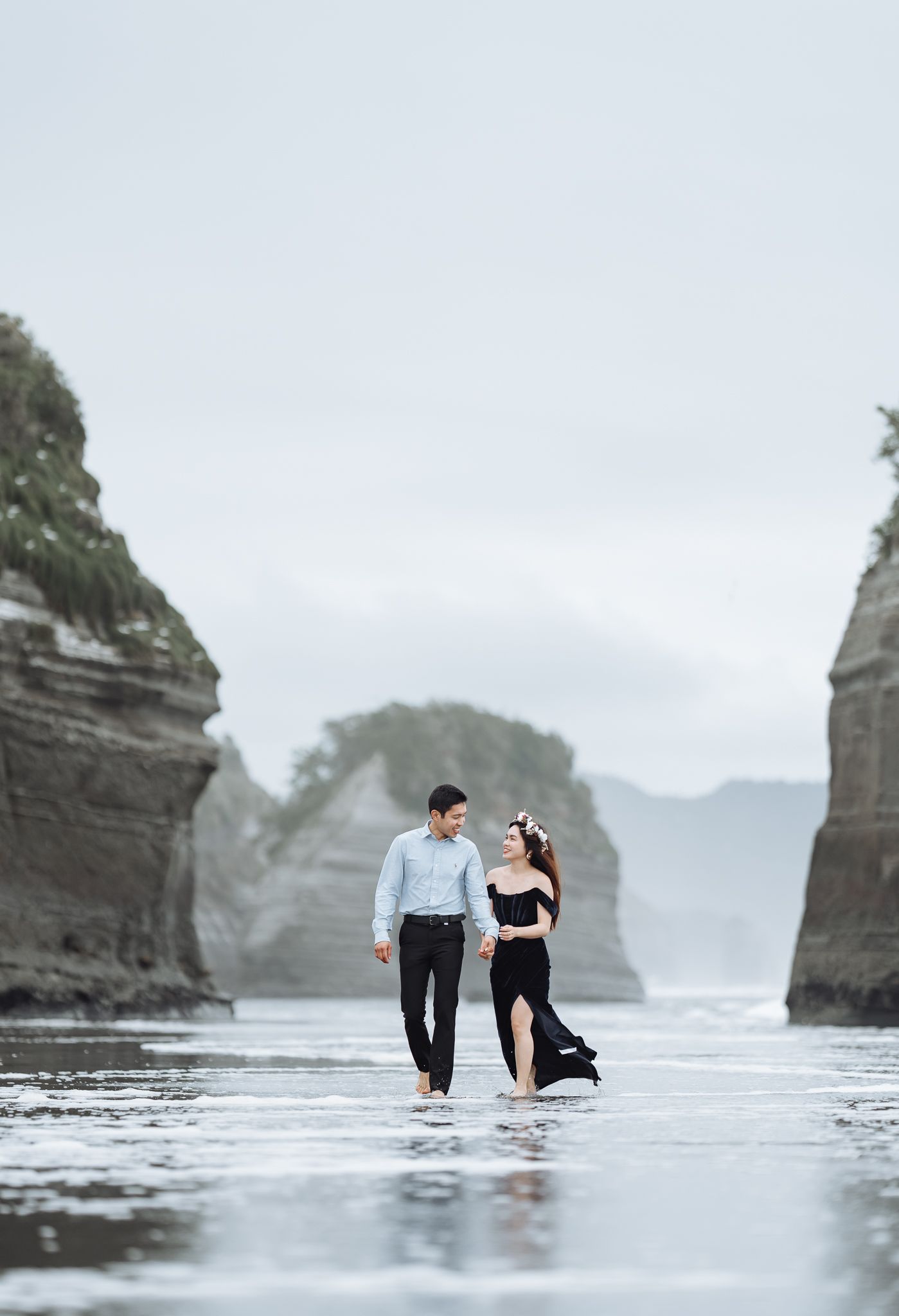Two people stand in shallow water near dramatic coastal rock formations.