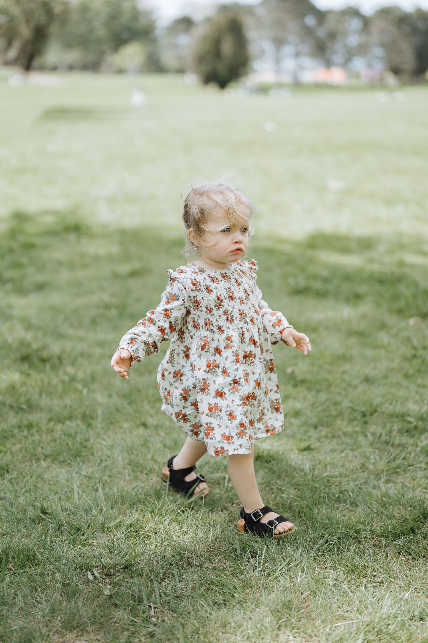 A toddler in a floral dress takes steps across a grassy field.