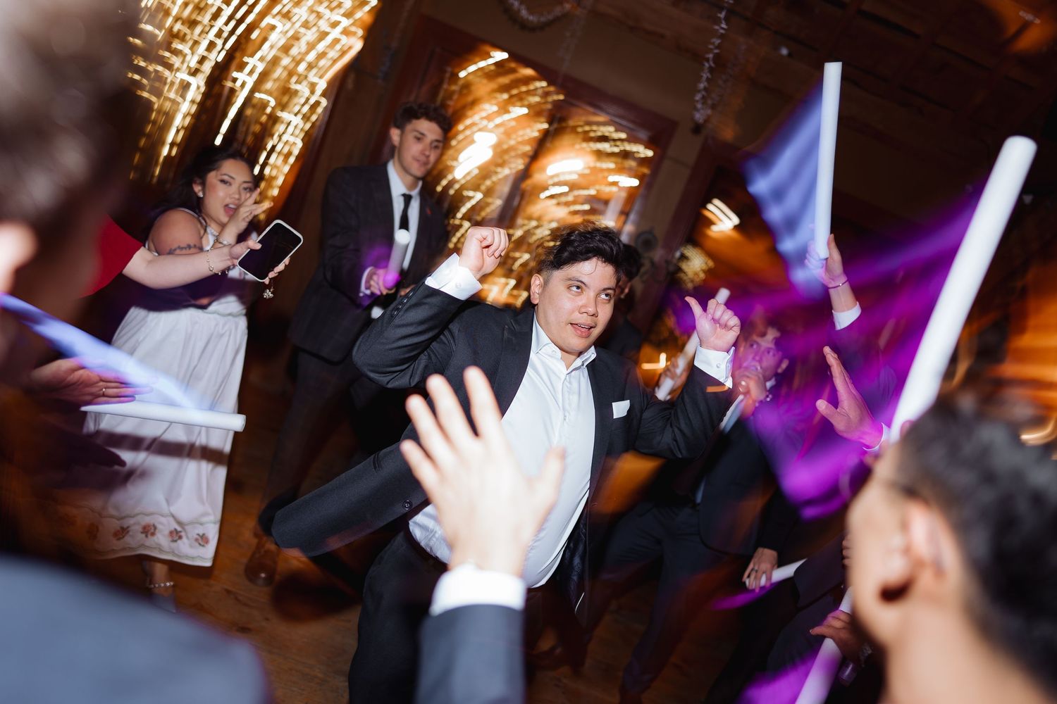 Wedding reception guests dance energetically on a dimly lit dance floor with colorful purple lighting effects.