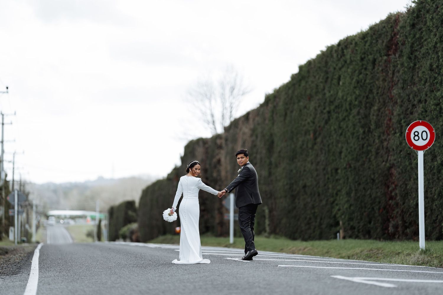 A couple holds hands while standing on a road with tall hedges and a speed limit sign.