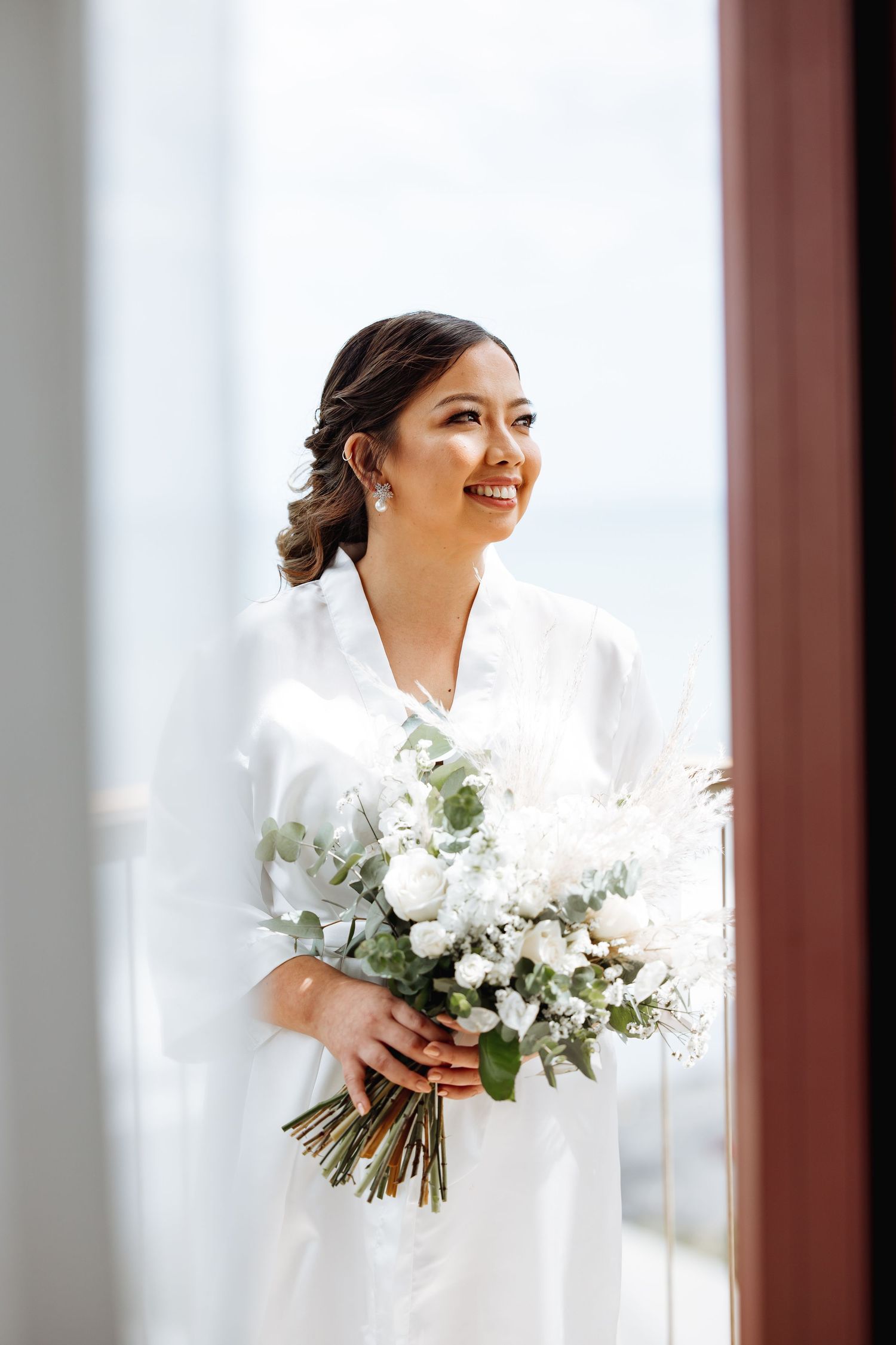 Bride in white dress holds elegant bouquet of white flowers against bright window.