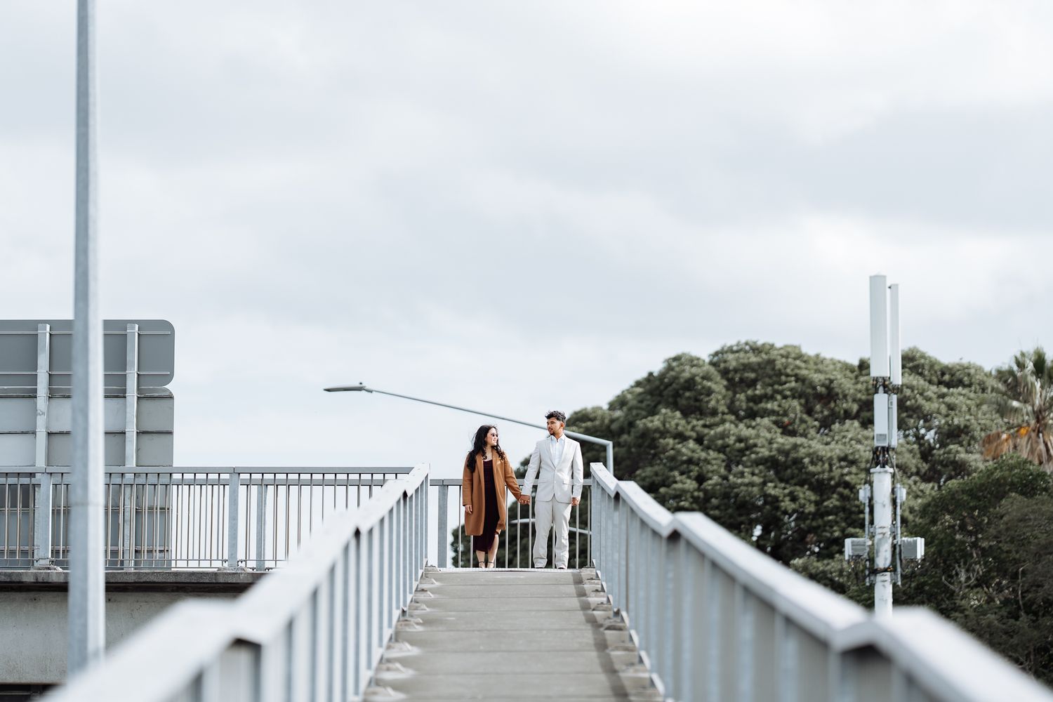 A couple walks along an elevated white railing walkway with trees and buildings in the background.