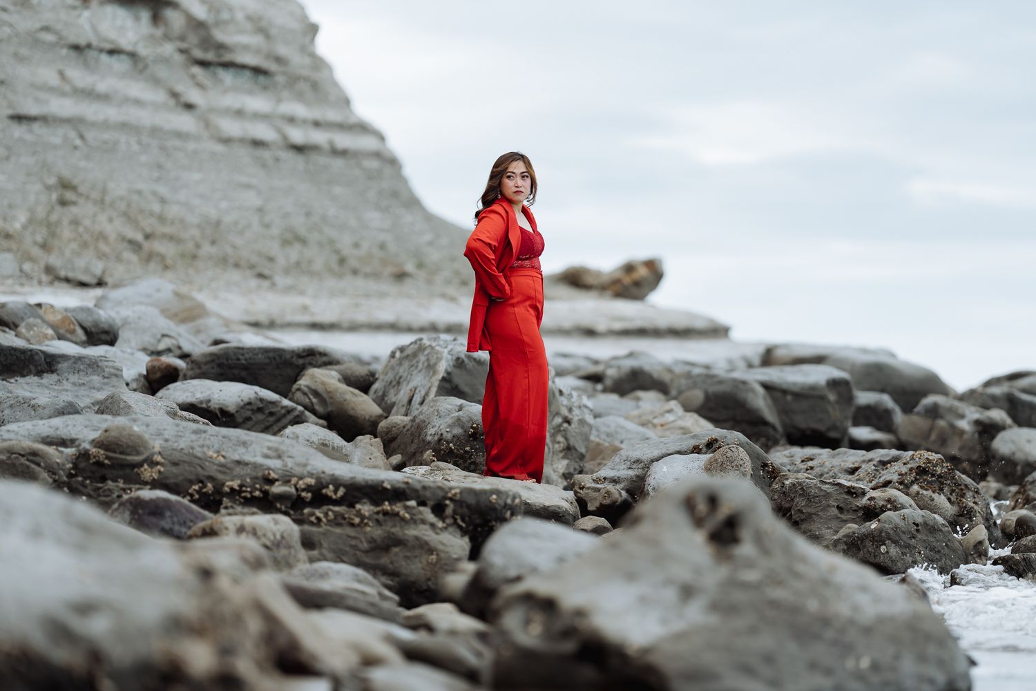 Dramatic coastal photography featuring small figure in red against rocky shoreline and cloudy sky.