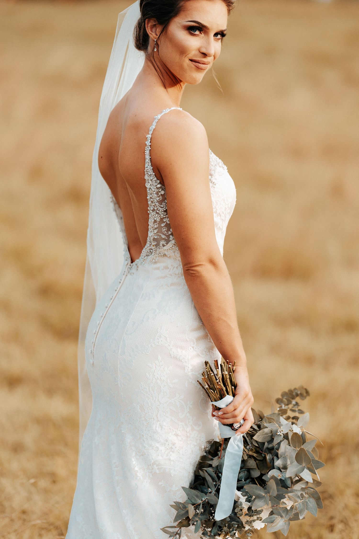Elegant backlit bridal portrait showcasing detailed lace gown and bouquet in golden field.