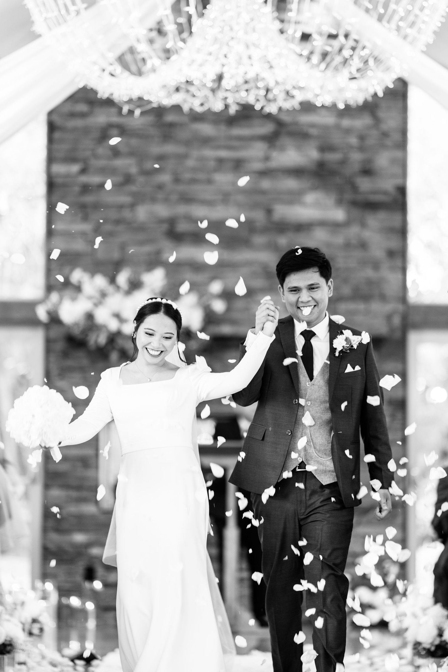 Black and white photo of joyful newlyweds walking through shower of flower petals.