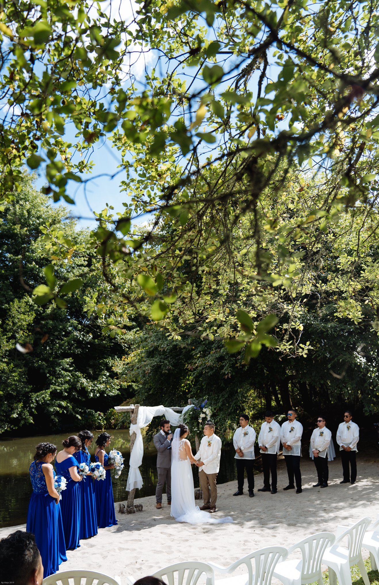 Outdoor wedding ceremony setup with blue-clad bridesmaids and groomsmen under green trees.