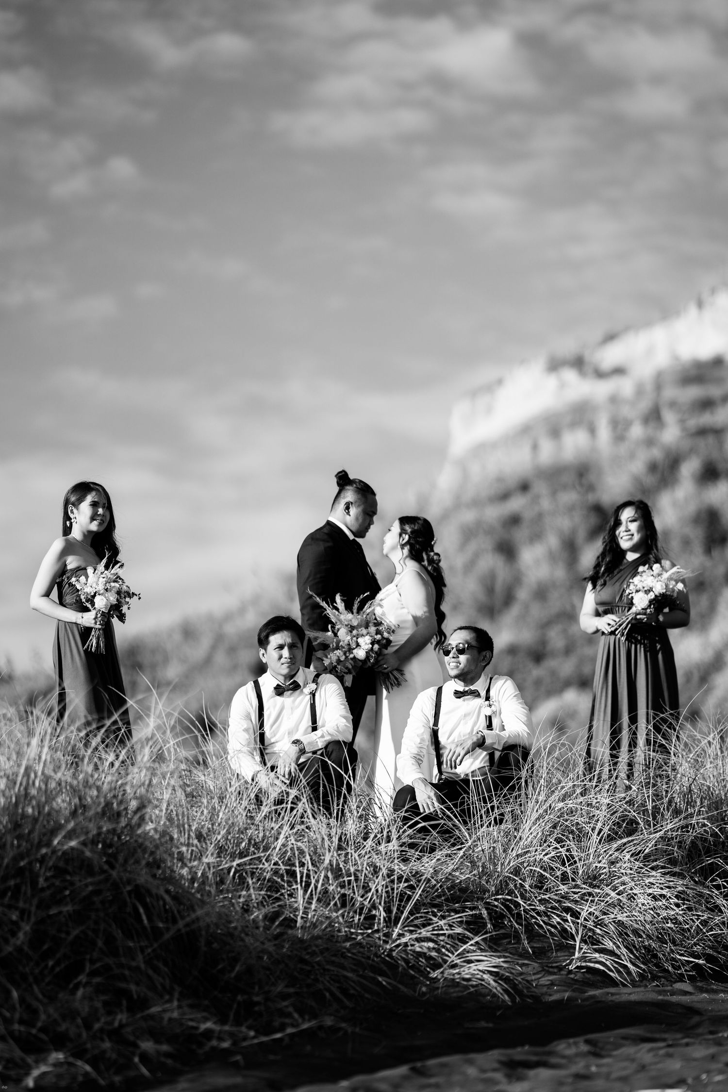 Black and white wedding party portrait with bridesmaids and groomsmen posing in tall grass with mountain backdrop.