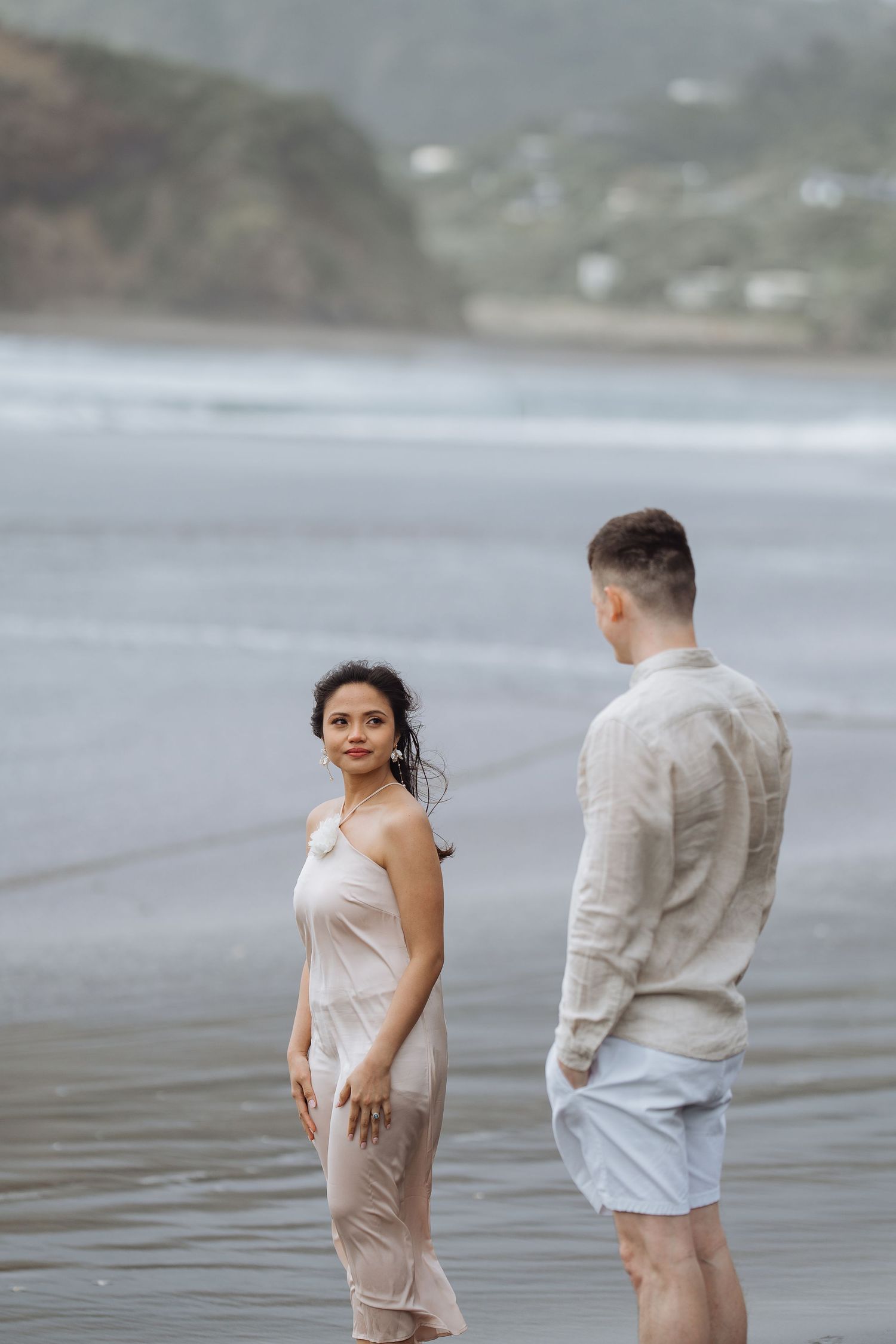 A couple stands together in shallow ocean water with misty cliffs in the background.