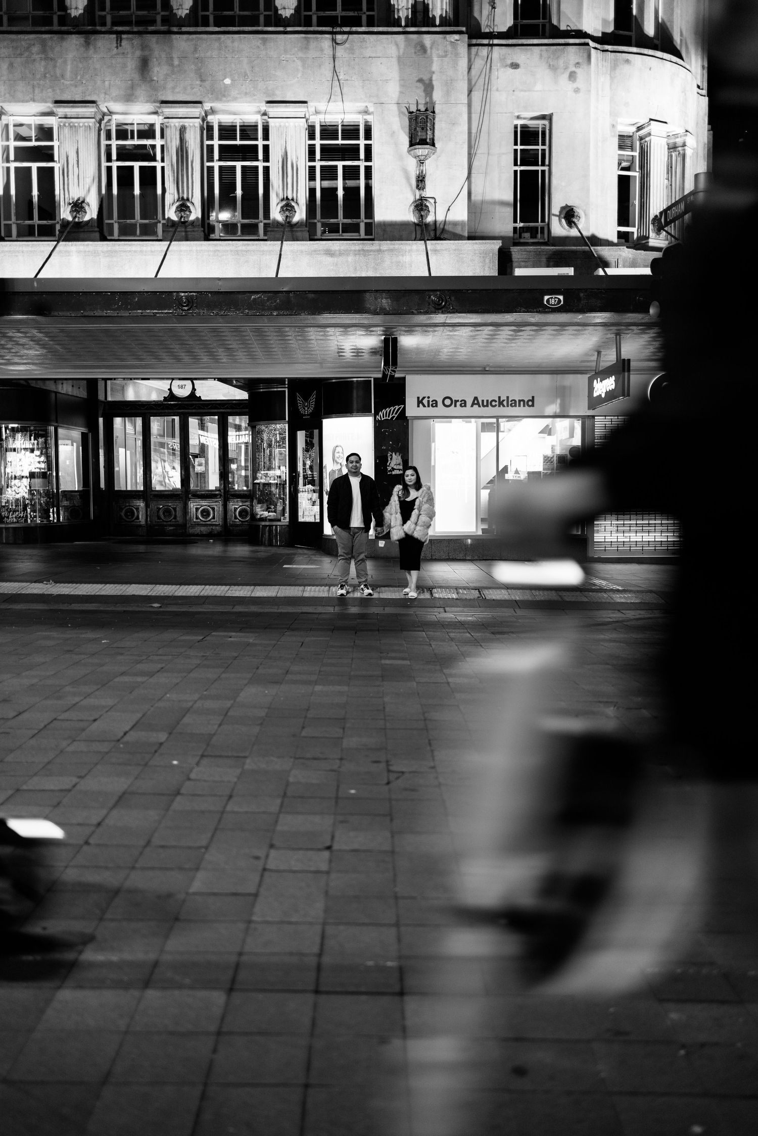 A moody nighttime street scene outside a lit storefront in black and white.