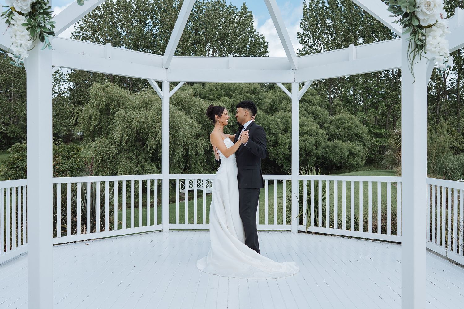 Wedding ceremony setup with white gazebo and railings surrounded by trees.