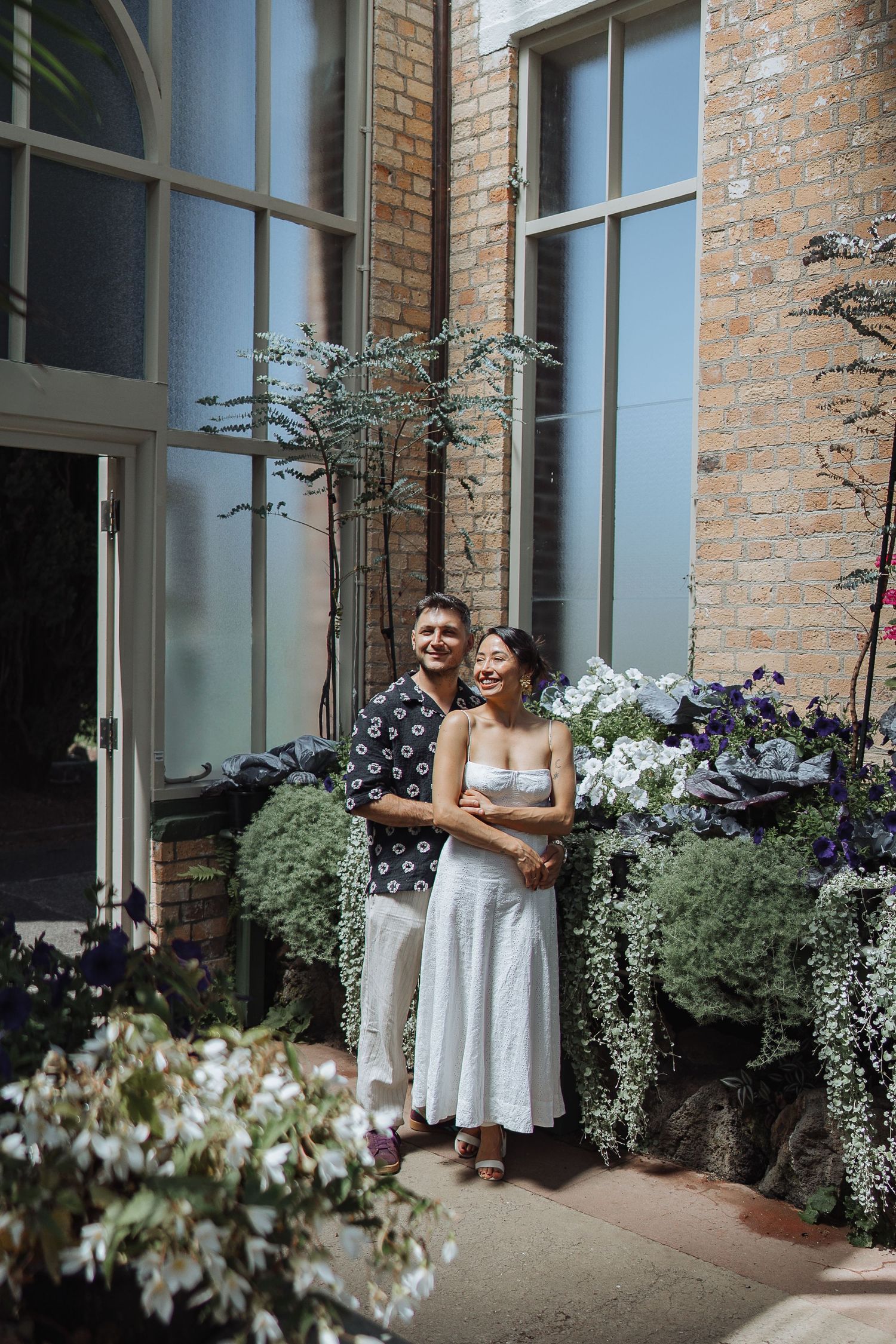 A couple embraces near a brick building with white flowers and greenery in the background.