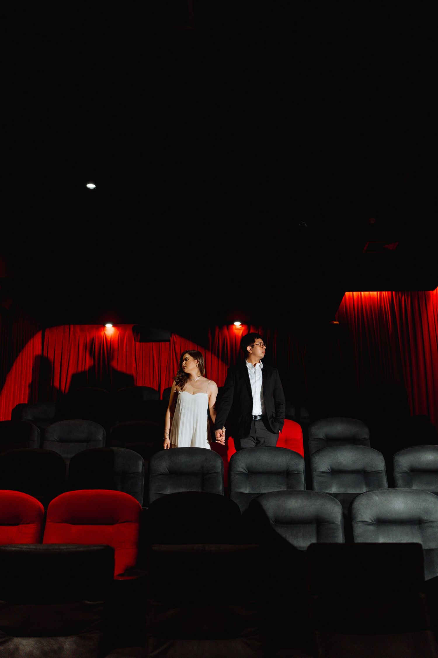 Couple standing in theater seats with dramatic lighting and red curtains.