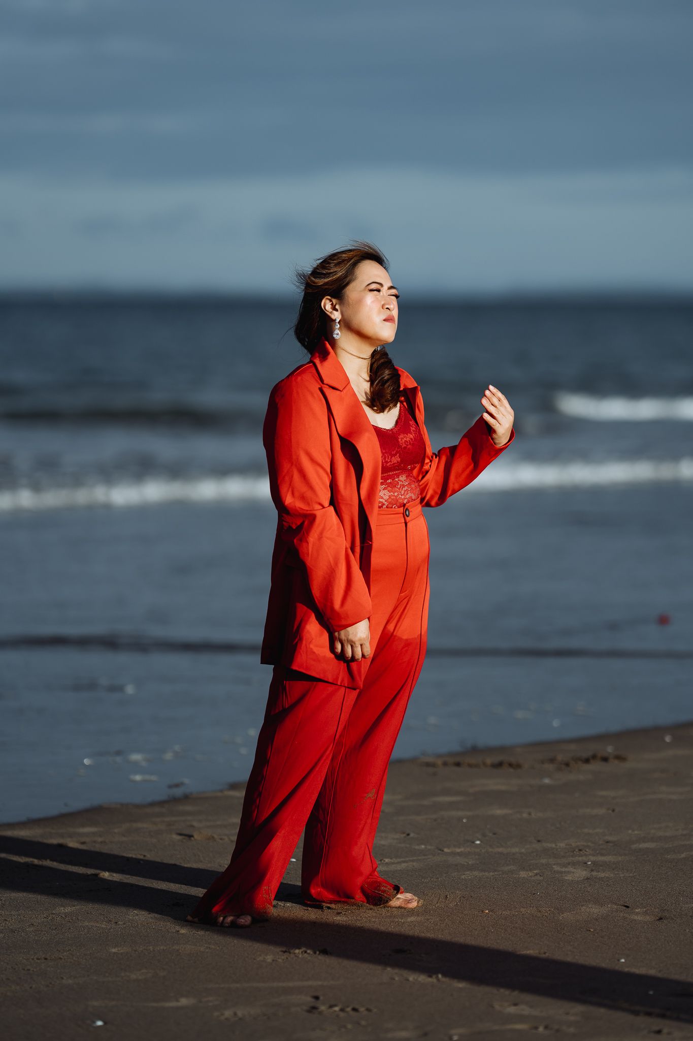 Figure in bright red outfit poses expressively on beach during sunset photoshoot.