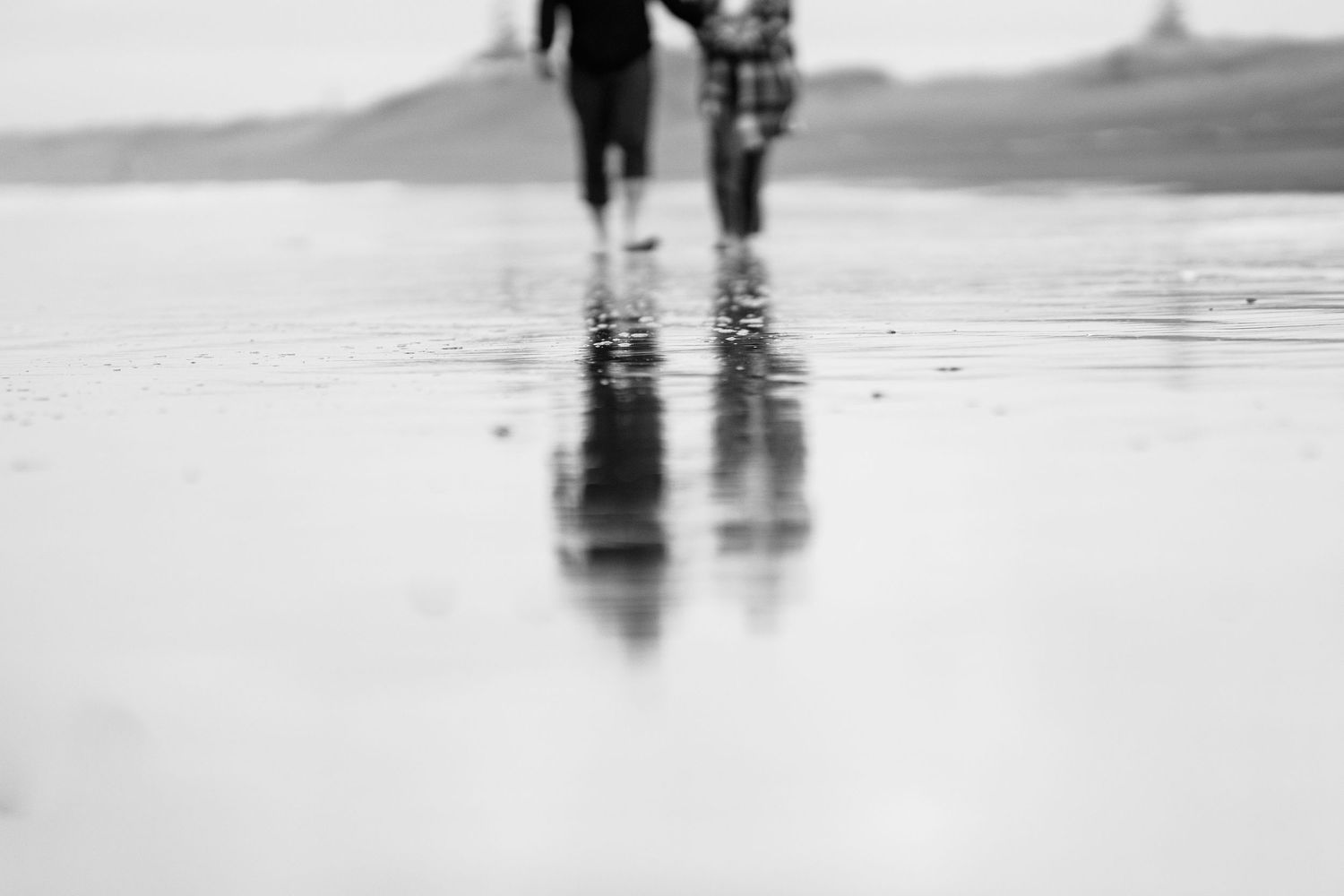 Black and white photo of two people walking on a wet reflective beach, their silhouettes mirrored on the sand.