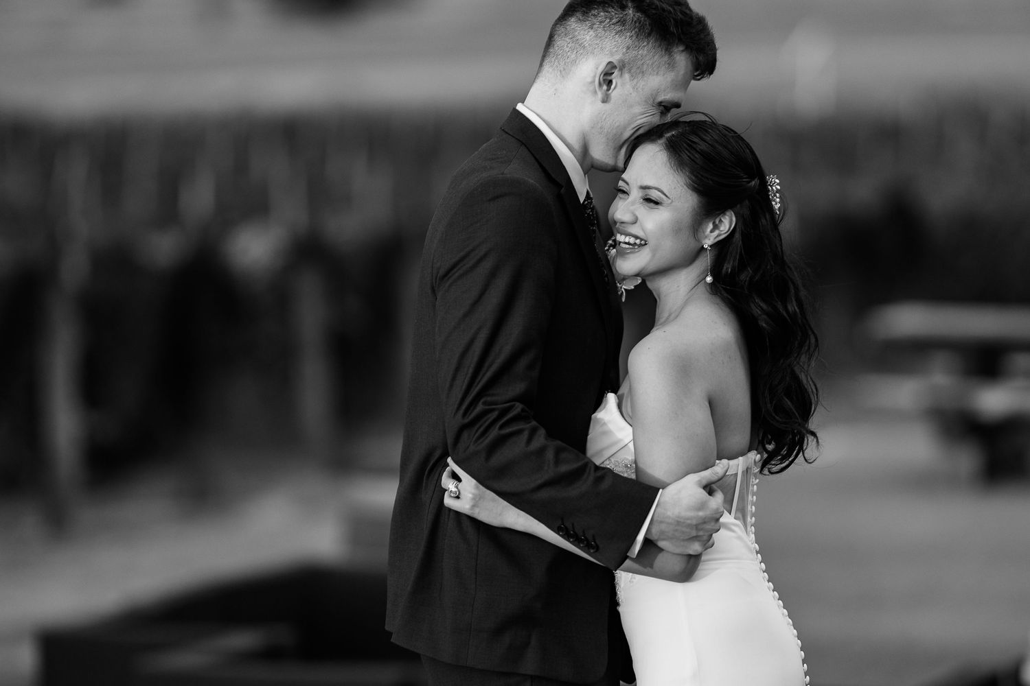 Black and white photo of a smiling bride in a white gown embracing a groom in a dark suit outdoors.