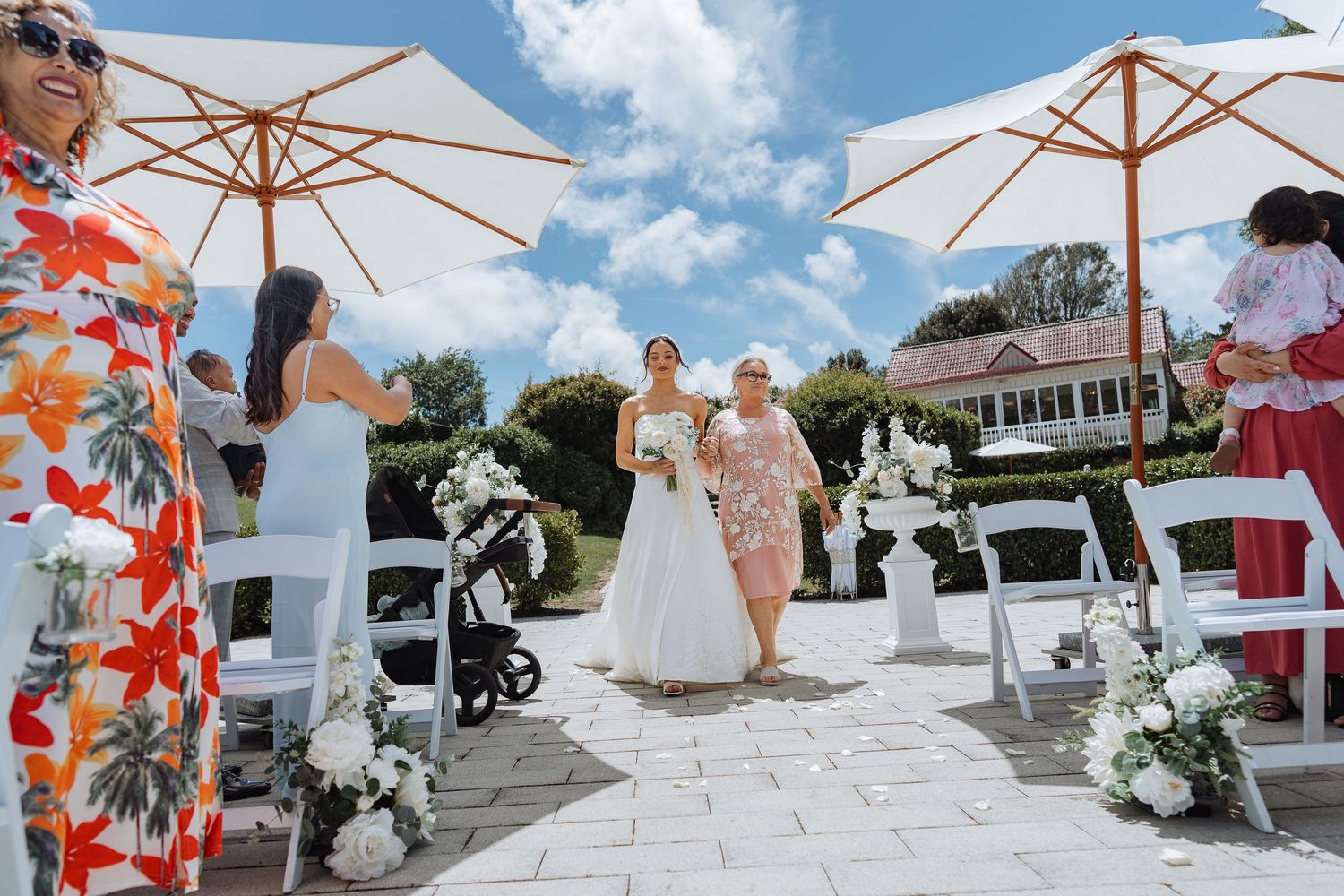Outdoor wedding venue with white umbrellas and floral decorations under blue summer sky.
