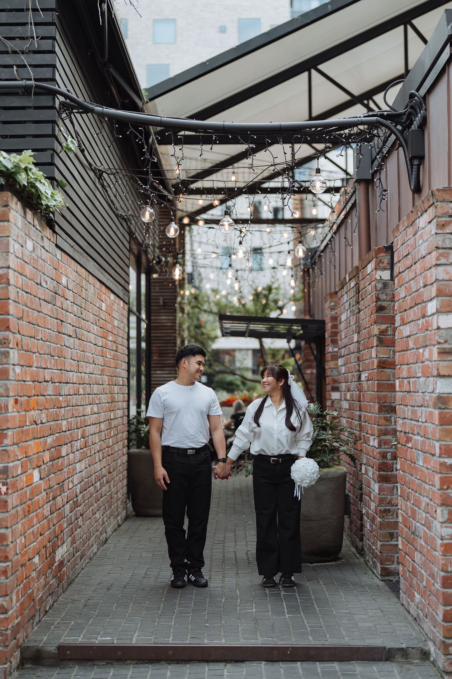 Romantic couple walking through a brick-lined covered walkway with string lights overhead.