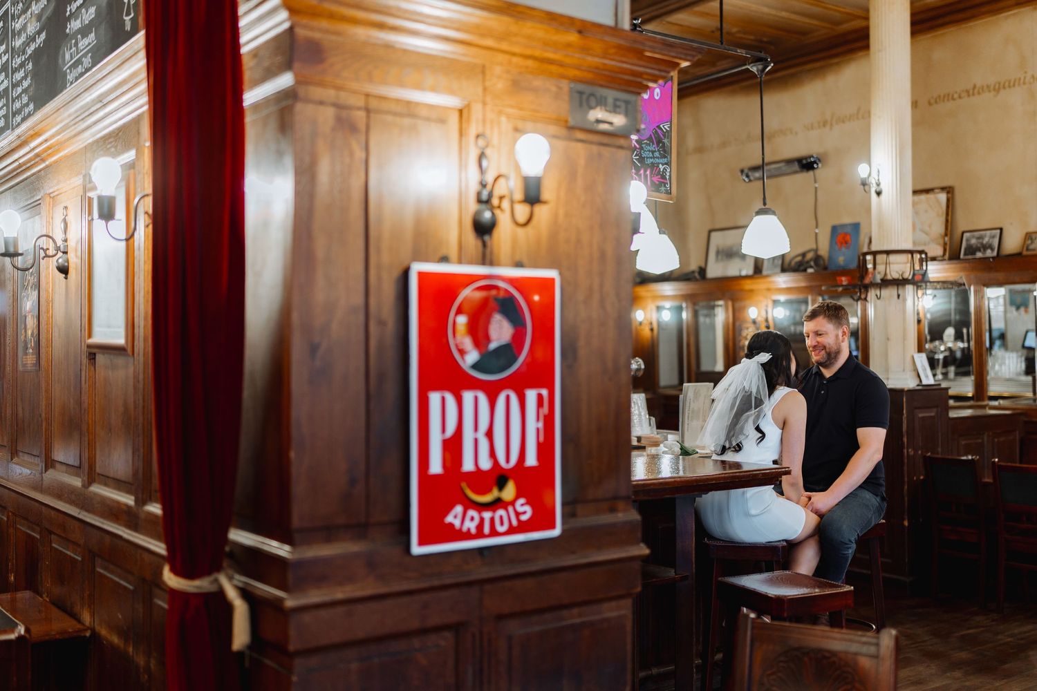A couple sits at a wooden bar with vintage decor and a Prof beer sign on the wall.