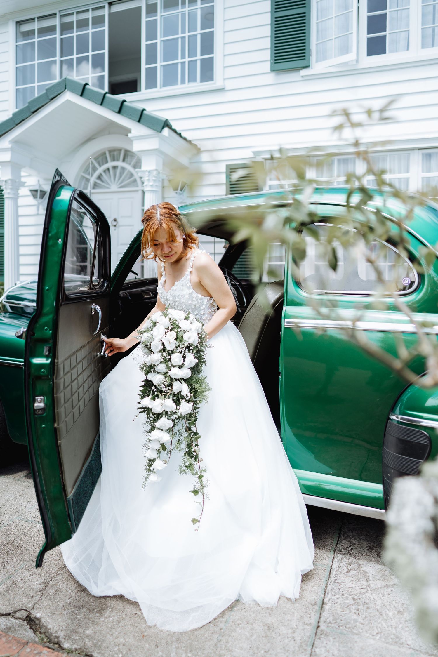 Bride in white gown entering vintage green car outside white colonial style house.