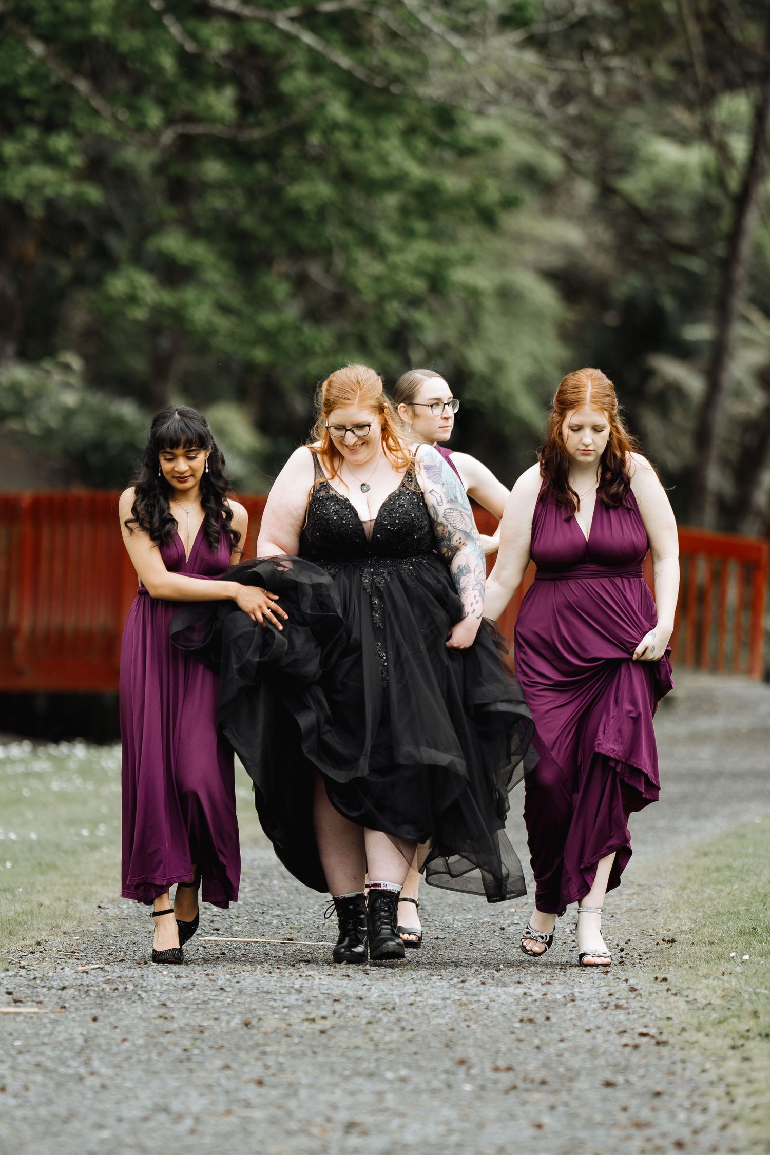 Bridesmaids in purple dresses and black shawls pose playfully on a gravel path.