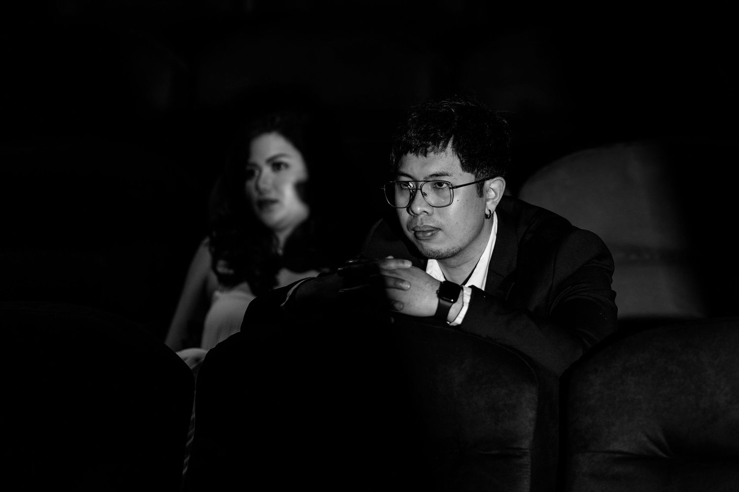 A couple sits together in darkness watching something intently.