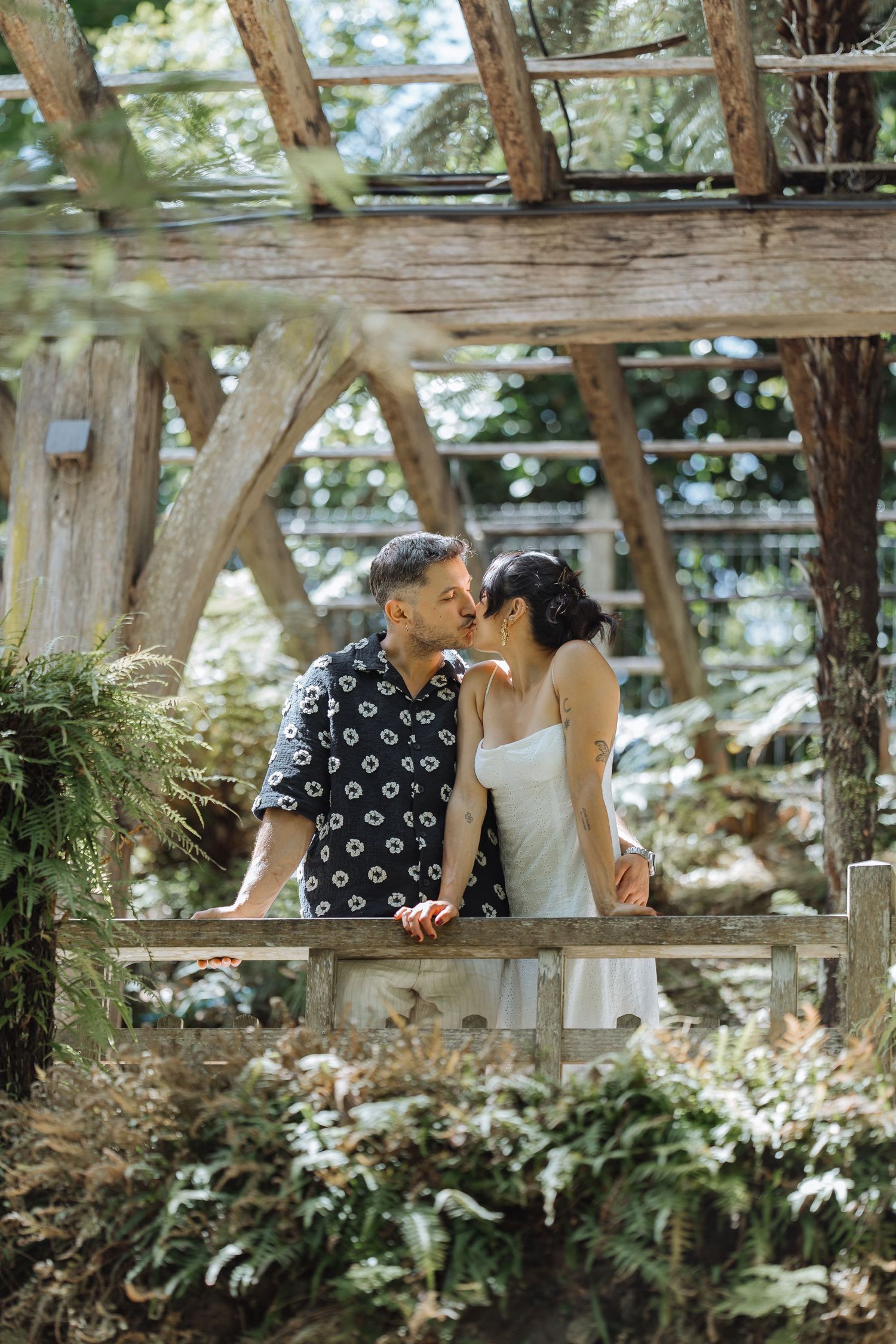 Couple sharing intimate moment under rustic wooden pergola surrounded by ferns.