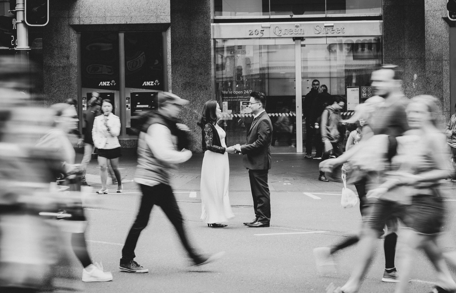 Black and white photo of a couple sharing a romantic moment on a busy city street with blurred pedestrians passing by.