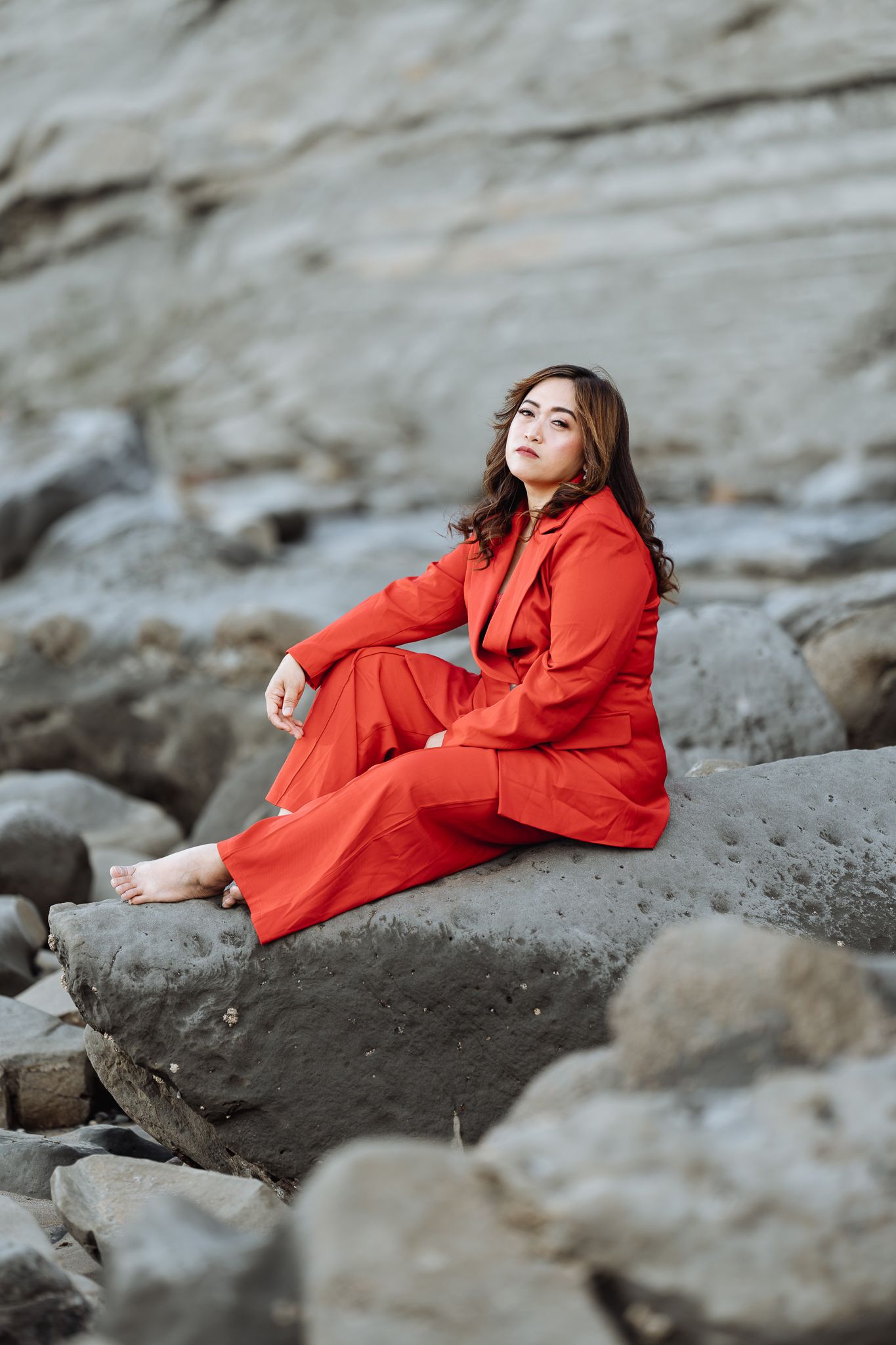 Figure in vibrant red dress poses dramatically on rocky coastal terrain.